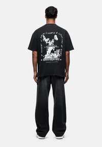 Black oversized t-shirt featuring white graphic print on the back. The design includes motifs of butterflies and a figure, text along the edges.