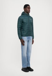 Grøn pufferjakke med hætte, med lynlås foran, quiltet tekstur og logo på brystet. Båret med lyseblå jeans og sorte sko.