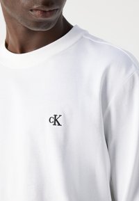 Hvit bomull langermet t-skjorte med en liten svart brodert "cK" logo på venstre brystområde. Glatt tekstur og crew neck-design.