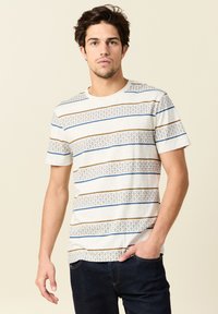 Vaaleanbeige lyhythihainen T-paita, jossa on vaakasuoria ruskeita ja sinisiä raitoja. Paidassa on kuviollinen yksityiskohta alareunassa.