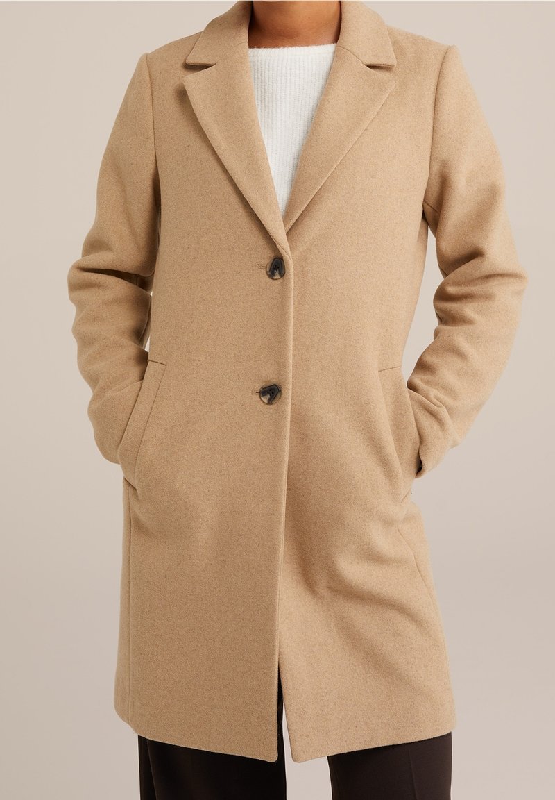 Beige villasekoitepäällystakki, jossa on lovikaulus, kaksi etutaskua, kolme nappia ja jäsennelty muotoilu. Pehmeä tekstuuri ja keskipitkä pituus.