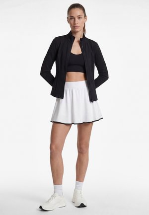 Vrouw staand met handen achter de rug, draagt een zwart kort jasje, een zwarte sport-bh, een witte rok met zwarte rand, witte sokken en witte sneakers.
