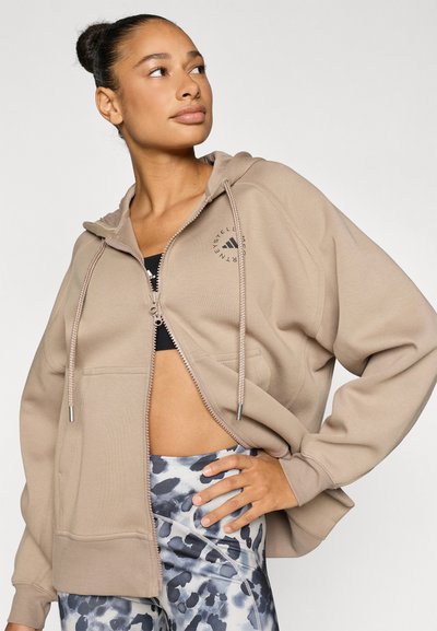 Felpa beige con zip e cappuccio con coulisse, decorata con un logo sul petto e due tasche frontali, abbinata a leggings con motivo a fantasia.