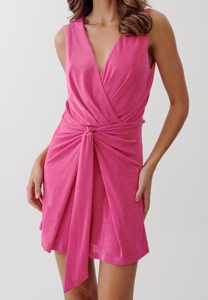 Femme portant une robe portefeuille sans manches rose vif avec un profond décolleté en V et un nœud à l'avant, se tenant devant un fond clair uni.