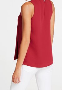 Blusa rossa senza maniche con colletto alto, texture liscia e spacchi laterali, abbinata a pantaloni bianchi aderenti. Design minimalista.