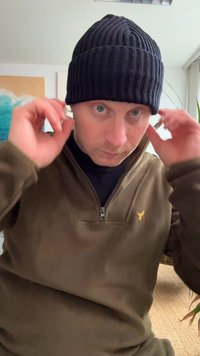 Černá žebrovaná čepice nošená s hnědou fleecovou bundou na zip. Vnitřní prostředí s rostlinami a abstraktním uměním v pozadí.