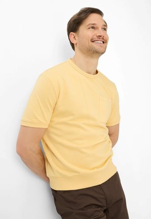 Homme appuyé contre un mur blanc, souriant et regardant vers le haut, portant une chemise à manches courtes jaune clair et un pantalon marron foncé.