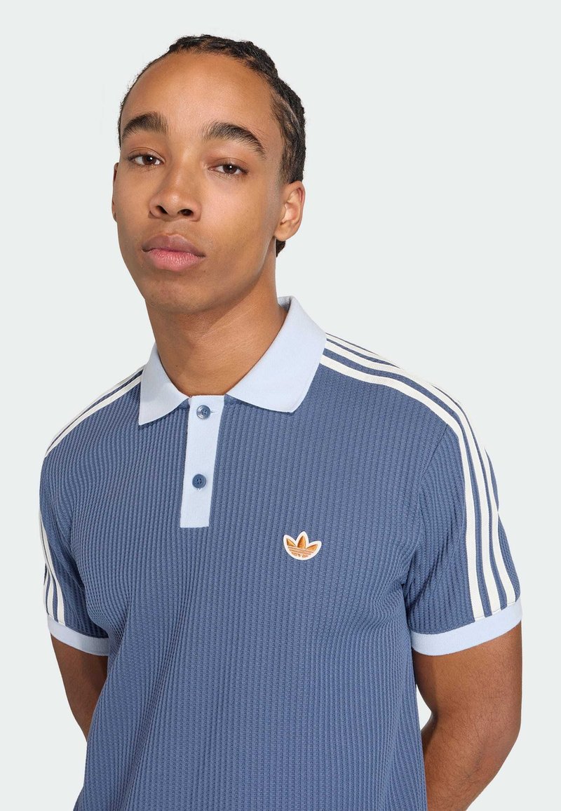 Giovane uomo con capelli intrecciati che indossa una polo Adidas blu con strisce bianche e logo, in piedi contro uno sfondo chiaro uniforme.