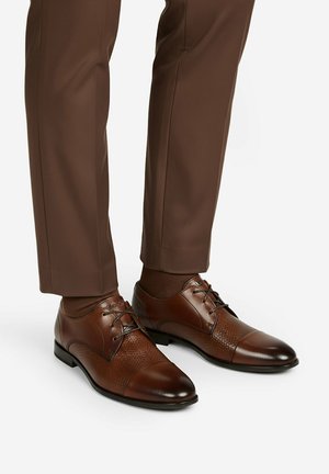 Brązowe skórzane eleganckie buty z teksturowanymi panelami i sznurowadłami, noszone z dopasowanymi brązowymi spodniami i skarpetkami na białym tle.