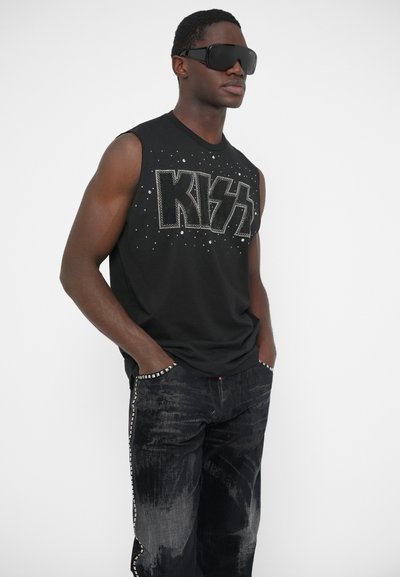 Camiseta negra sin mangas con el logo "KISS" de pedrería, combinada con jeans negros desgastados que presentan detalles laterales con cuentas. La modelo lleva gafas de sol de gran tamaño.