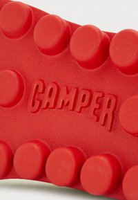 Sole de borracha vermelha com saliências redondas e texturizadas. O nome da marca "CAMPER" está estampado em vermelho contrastante. Acabamento liso e mate.