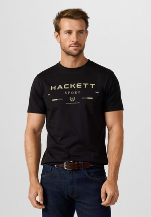 Man draagt een zwart Hackett Sport T-shirt en donkerblauwe spijkerbroek met een bruine riem, staand tegen een effen lichtgrijze achtergrond.