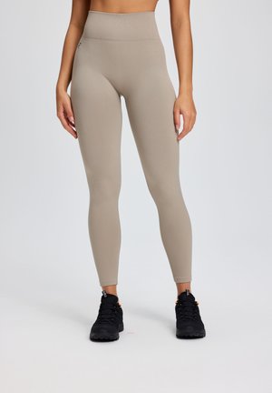 Kvinde iført beige leggings med høj talje og sorte træningssko, stående mod en ensfarvet lys baggrund.
