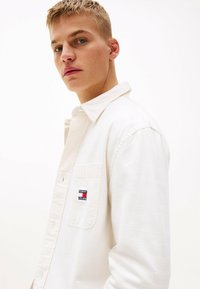 Biała koszula z długim rękawem i kołnierzykiem zapinanym na guziki, z kieszonką na piersi oraz małym haftowanym logo Tommy Jeans. Gładka tekstura materiału.