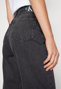 Jeans de mezclilla negros de talle alto con dos bolsillos traseros. Presentan un parche con el logo en la cinturilla y un corte recto con una textura sutil.