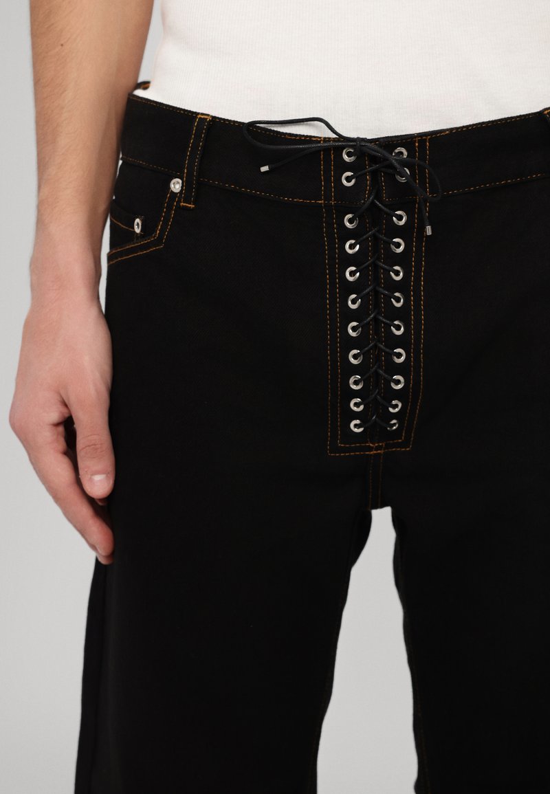 Black denim pants featuring a unique lace-up front with metal eyelets, contrasting orange stitching, and standard side pockets.
