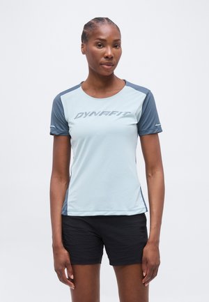 Γυναίκα όρθια, φοράει ένα κοντομάνικο Dynafit σε ανοιχτό μπλε και γκρι και μαύρο σορτς, σε απλό λευκό φόντο.