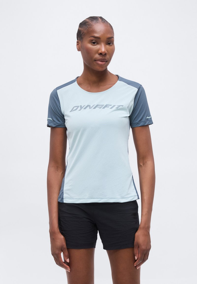 Γυναίκα όρθια, φοράει ένα κοντομάνικο Dynafit σε ανοιχτό μπλε και γκρι και μαύρο σορτς, σε απλό λευκό φόντο.