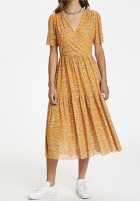 Mujer vestida con un vestido envolvente floral amarilla mostaza con mangas cortas y zapatillas blancas, de pie frente a un fondo blanco liso.