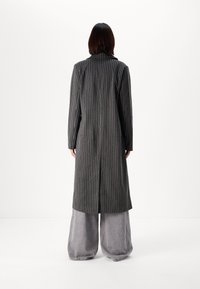 Dlouhý šedý kabát s proužky a rozparkem vzadu, se sklopeným límcem a dlouhými rukávy, doplněný volnými šedými kalhotami.