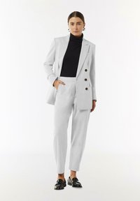 Lichtgrijs op maat gemaakt pak met een dubbele knoop blazer, een enkele zak en tapered broek. Gekocht over een zwarte coltrui, aangevuld met glanzende zwarte loafers.