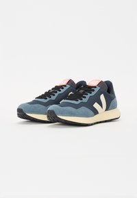 Sneaker in suede blu e navy con lacci neri, dettagli bianchi, una linguetta rosa e pannelli testurizzati su una suola in gomma nera.
