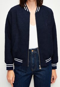 Granatowa kurtka bomberka z teksturowanej tkaniny, białe prążkowane mankiety oraz zamek błyskawiczny z przodu. Haftowane logo na piersi.