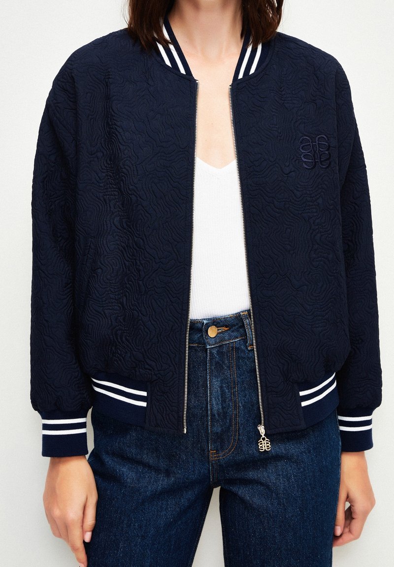 Granatowa kurtka bomberka z teksturowanej tkaniny, białe prążkowane mankiety oraz zamek błyskawiczny z przodu. Haftowane logo na piersi.