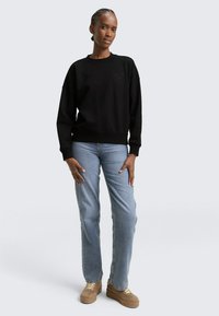 Donna in piedi che indossa una felpa nera, jeans azzurri chiari e sneakers beige con zeppa su uno sfondo chiaro e semplice.