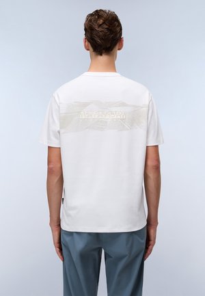 Mand iført en hvid kortærmet t-shirt med lys grafik og teksten "NAPAPIJRI" øverst på ryggen, kombineret med blå bukser.
