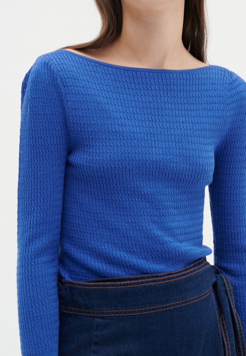 Mujer vestida con una blusa de manga larga azul texturizada metida dentro de una falda azul oscura con costuras naranjas visibles.