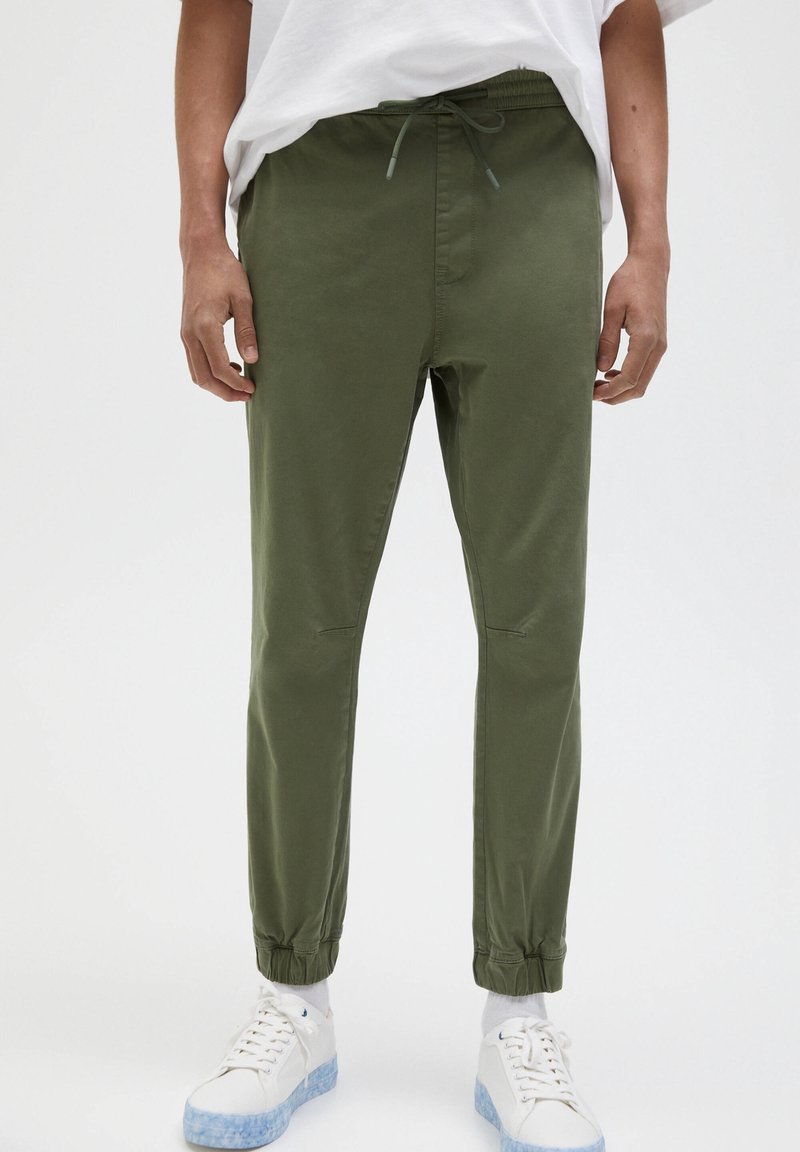 Pantalones jogger verde oliva con puños elásticos y cintura con cordón, fabricados en un tejido suave con bolsillos laterales y detalles de costura en las rodillas.