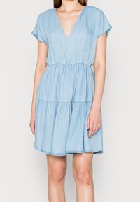 Vaaleansininen farkumekko, jossa on V-pääntie, lyhyet hihat ja joustava vyötärö, ja jonka helma on kerroksellinen ja liehuva, sekä pehmeä rakenne.