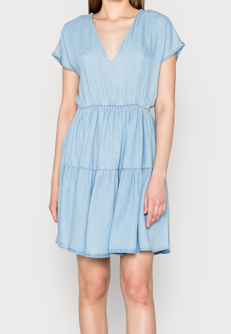 Vaaleansininen farkumekko, jossa on V-pääntie, lyhyet hihat ja joustava vyötärö, ja jonka helma on kerroksellinen ja liehuva, sekä pehmeä rakenne.