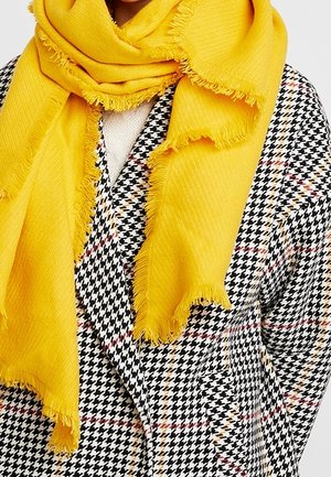 Écharpe jaune vif à franges drapée sur un manteau à motif pied-de-poule noir et blanc avec des lignes subtiles rouges et beiges.