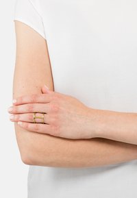 Bague en or avec double bande au texture lisse, présentant deux barres parallèles. La main repose contre un bras, tous deux légèrement bronzés.