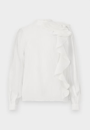 Blouse blanche à manches longues avec un col montant, présentant un détail de volants superposés sur une épaule et une texture lisse. Les poignets sont boutonnés.