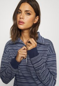 Navy blue zippered pullover with white geometric pattern, featuring a wide collar and ribbed cuffs. Soft fabric texture, casual design.