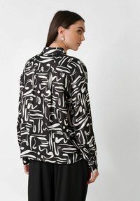 Woman with pearl earrings wearing a black shirt with white abstract brushstroke patterns, paired with black pants, facing sideways.