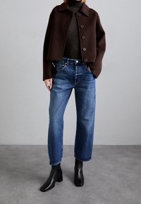 Brun kort jacka med stora knappar, mörk tröja, blå raka jeans och svarta ankelstövlar. Enkelt utformning, mjuka texturer.