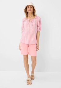 Helles, rosa gestreiftes Blusenshirt mit Schlüssel-Ausschnitt und Puffärmeln, kombiniert mit hellrosa Shorts. Das Modell trägt einen weißen Bucket Hat und Sandalen.