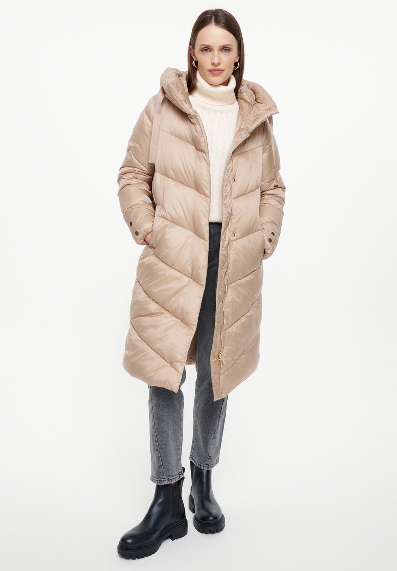 Beige pufferjack met capuchon, diamantpatroon en drukknopen. Draag het over een crèmekleurige turtleneck, gecombineerd met grijze jeans en zwarte laarzen.