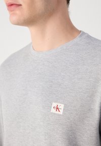 Gros plan sur une personne portant un pull texturé gris clair avec un patch logo Calvin Klein rouge et blanc sur la poitrine.