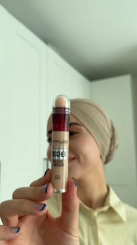 Una donna con un foulard beige tiene il correttore multiuso Maybelline Instant Th er Lösch er 03 vicino al viso, con unghie con punte blu visibili.