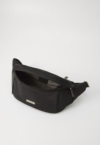Zwarte nylon heuptas met een gladde textuur, ritssluiting, zilveren logo plaat en verstelbare schouderband. Binnenin is de tas gevoerd met een lichtere tint.