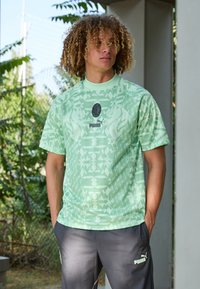 Camisa atlética de color verde claro con patrones geométricos, de manga corta y cuello redondo. Logotipo negro de Puma en el pecho y emblema del club en el centro.