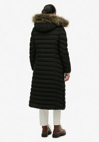Cappotto puffo nero con cappuccio in pelliccia sintetica, con sezioni trapuntate verticali, design lungo fino al ginocchio, indossato con pantaloni beige e stivali marroni.