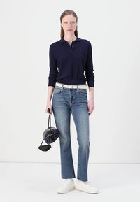 Polo de manga longa de malha navy, jeans de perna reta azuis claros, ténis brancos e uma mochila preta com cordão. Conjunto simples e casual.