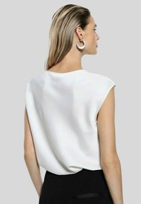 Blusa bianca senza maniche con una texture liscia e scollatura rotonda, abbinata a un fondo nero, su uno sfondo grigio.