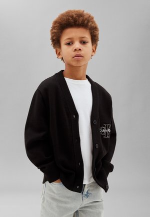 Mladý chlapec s kučeravými vlasmi, oblečený v čiernom kardigáne Calvin Klein cez bielu košeľu a svetlomodré džínsy, stojaci pred sivým pozadím.